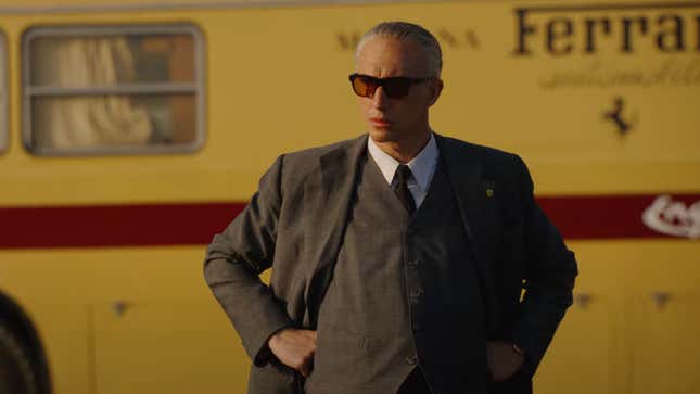 A man in a brown three-piece suit and dark sunglasses stands with his hands on his hips in front of a Ferrari yellow transporter with a red stripe