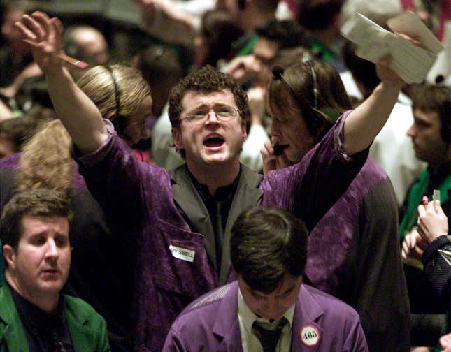 A man in glasses and a purple coat stands on a trading floor, shouting , with his arms raised. 