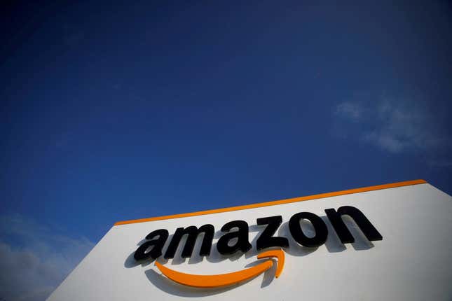 An image of the Amazon logo on the facade of a building, taken from the ground looking up. The sky in the background is blue with a couple wispy clouds.