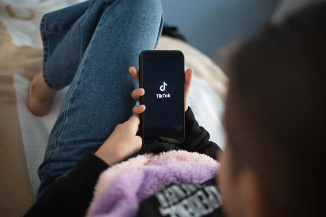 A young woman sits back on the couch holding a phone that has the TikTok logo on it.