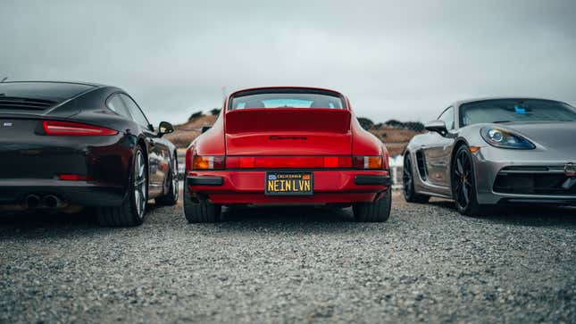 A red 1980s Porsche 911 Carrera with a ducktail spoiler is parked on gravel. Rear view.