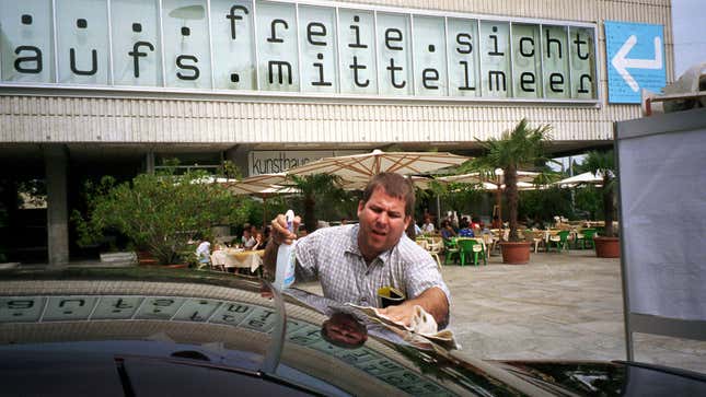 A photo of a person spraying the windshield of a car outside a museum. 