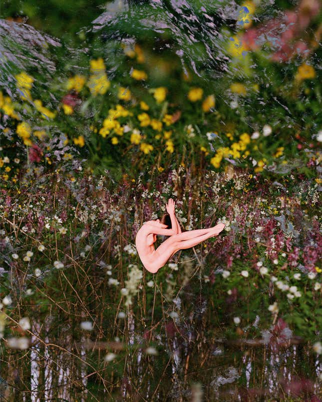 A naked woman folds her body inward against a backdrop of flowers.