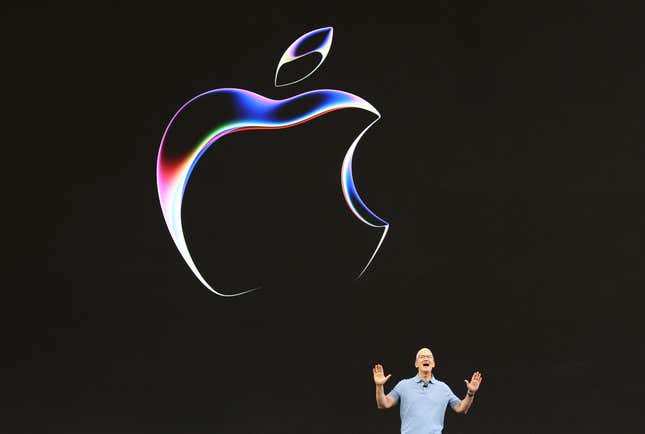 Tim Cook is small in the bottom right side of the photo speaking with both of his hands up, an apple logo on a black backdrop is behind him