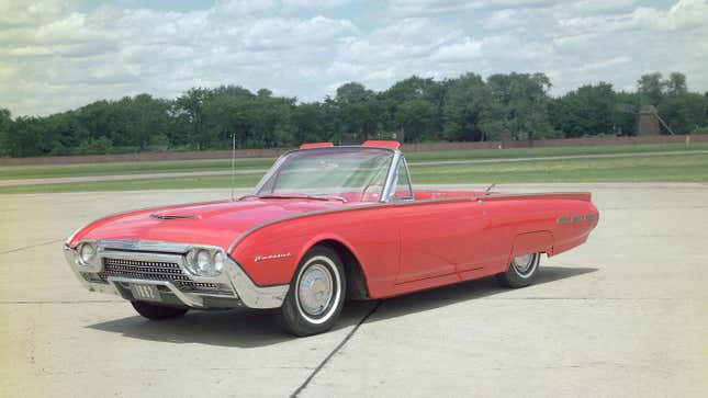 A red 1962 Ford Thunderbird parked in a wide open paved lot with trees and blue sky in the background