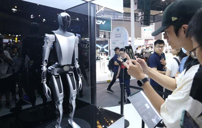 gray and silve Tesla Optimus robot on display in a glass case. A crowd of people are taking pictures with their phones