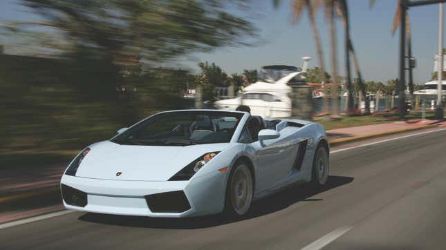 A light blue Gallardo Spyder driving past some docks with the roof down