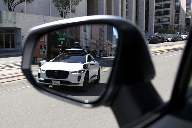 A Waymo autonomous vehicle is seen reflected in a car wing mirror while driving on the road.