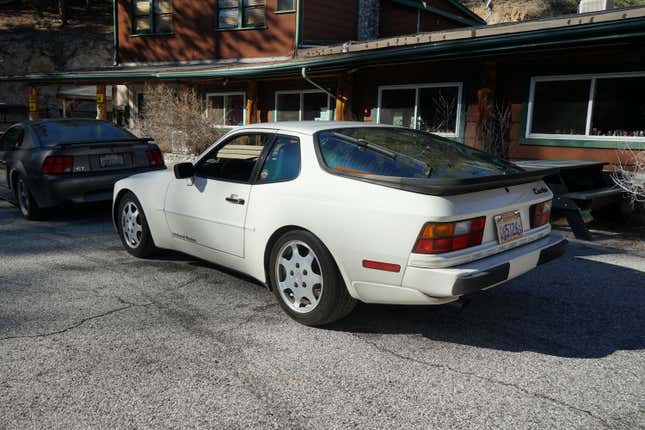 A white Porsche 944 Turbo is parked behind a gray Ford Mustang.