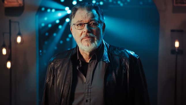 Actor Jonathan Frakes stands on a dimly-lit set, illuminated from behind by beams of light.