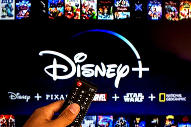 Sur cette photo illustrative on voit un gros plan d’une main tenant une télécommande de télévision affichée devant le logo Disney+.