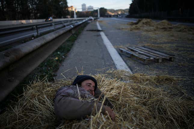 A farmer lies in a pile of hay after spending the night at a highway barricade in Aix-en-Provence, southern France, Tuesday, Jan. 30, 2024. France&#39;s protesting farmers encircle Paris with tractor barricades, vowing a &quot;siege&quot; over grievances. (AP Photo/Daniel Cole)