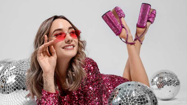 A woman wearing a pink sequined dress and pink sequined platform shoes lies on her stomach amongst disco balls. 