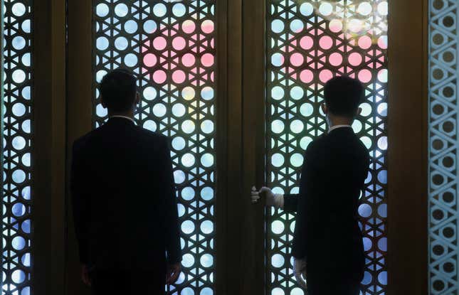 Two people wearing suits are shown from behind peering through a decorate grated door.