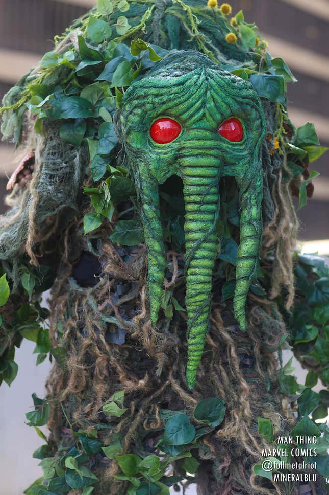 A cosplayer stands dressed as Man-Thing, complete with creepy red eyes and ivy twisting around their body.