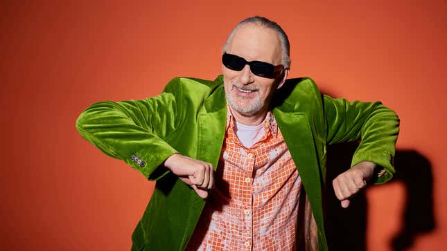 An older man wearing dark sunglasses, a green velvet blazer, and a pattern shirt dances in front of an orange backdrop. 