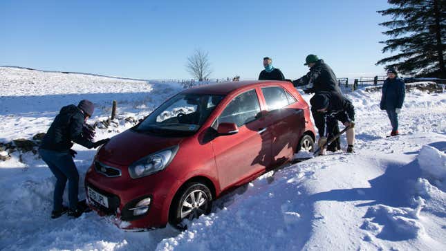 A photo of a red car stuck in a snow with people digging it out. 