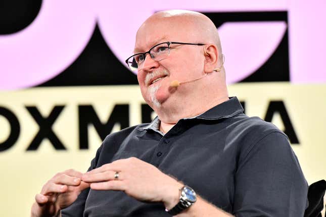 Kevin Scott speaking in front of a backdrop that includes pink and black letters on a black and yellow background