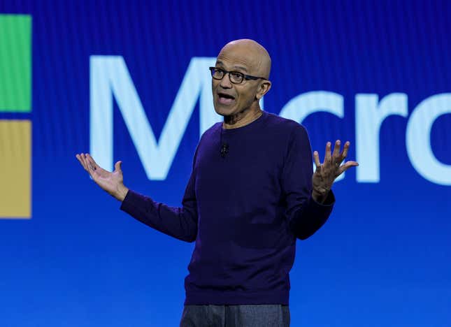 Satya Nadella standing in front of a Microsoft backdrop with both of his arms raised mid-speaking