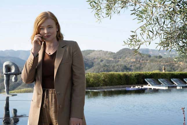 Succession character Shiv Roy is holding a phone to her ear while standing next to an outdoor pool lined with chairs. A view of green hills is in the background.