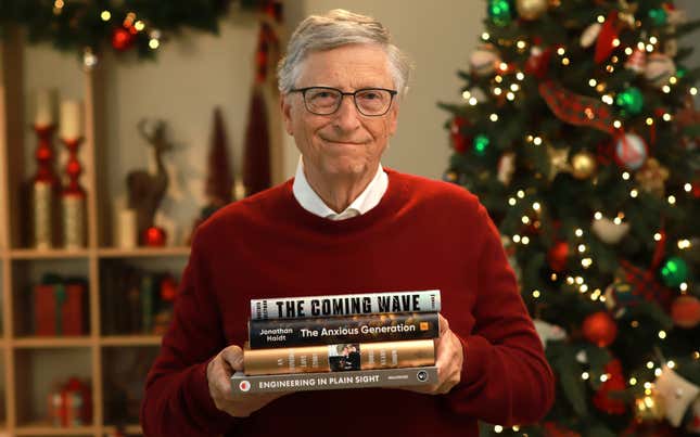 Bill Gates sosteniendo cuatro libros, vistiendo un suéter rojo y una camisa blanca abotonada frente a un árbol de Navidad