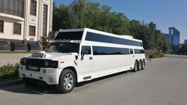 A white Hummer limousine parked on the side of the road with an extended roof and three rear axles