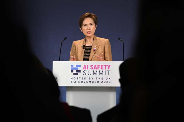 Josephine Teo wearing a tan blazer and black and tan shirt underneath speaking at a podium, framed by shadow outlines of people