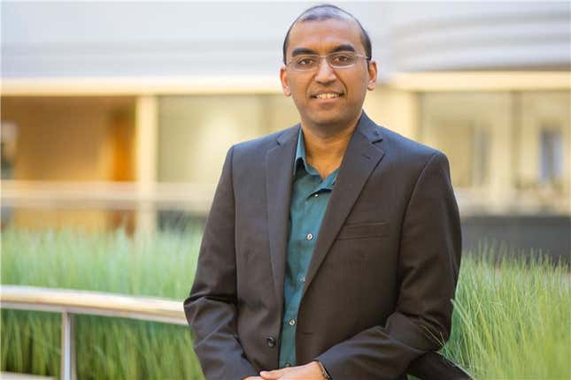 headshot of Arun wearing a suit jacket and blue button down standing on a bridge in front of grass