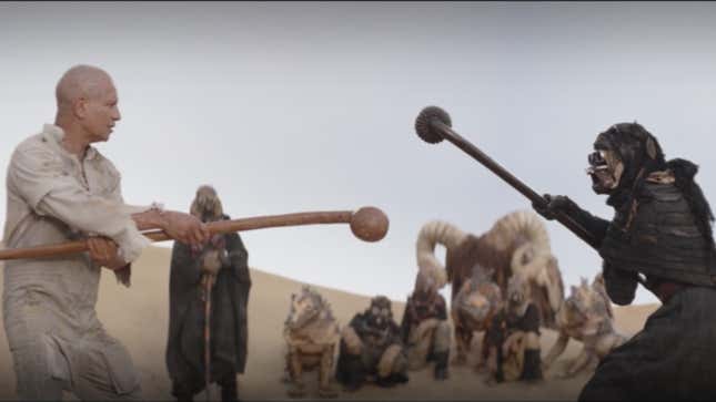 A Tusken clad in black teaches Boba Fett to wield his gaderffii stick as others watch on.