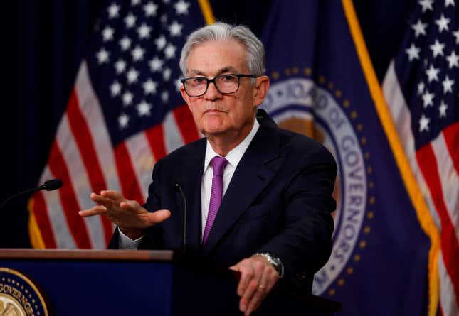 Federal Reserve chair Jerome Powell stands at a podium, one hand gripped on the side, the other gesturing, his face looking serious. US flags are in the background.