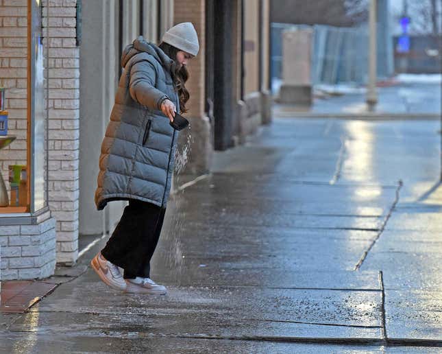 Mariah Sanders sprinkles rock salt to melt the ice covered sidewalk outside the Hi Honey salon downtown Bismarck, N.D., on Tuesday morning, Dec. 26, 2023, as ice covered streets and sidewalks made walking and driving difficult. Sanders said she was expecting a customer, but others had called in to postpone due to weather conditions. (Tom Stromme/The Bismarck Tribune via AP)