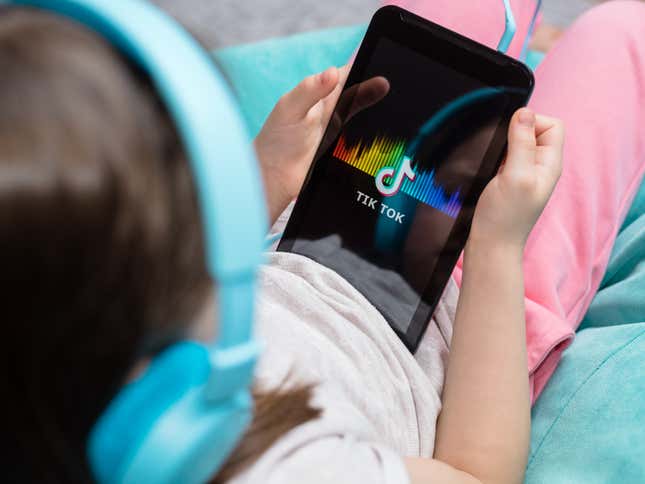 A young girl with blue headphones lays back on a chair cradling a tablet with the TikTok logo on it.