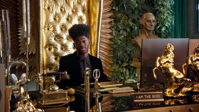 Lil Nas X sits at a golden desk at Riot Games' office. 
