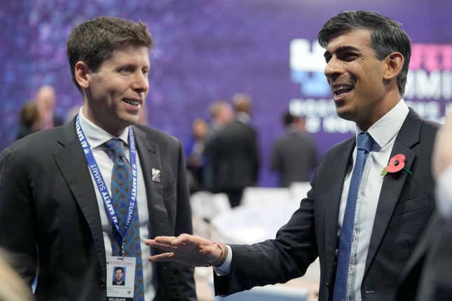 Sam Altman (left) and Rishi Sunak (right) both wearing suits and ties and speaking to each other, the background is blurred and has a purple backdrop and people