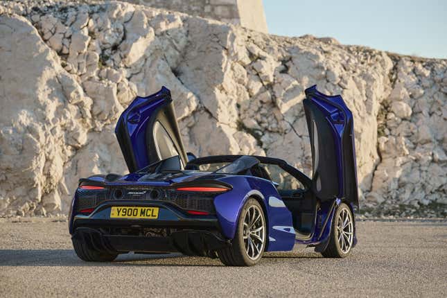 A rear 3/4 shot of the deep blue Artura Spider parked in front of some white rocks with its vertically opening doors open