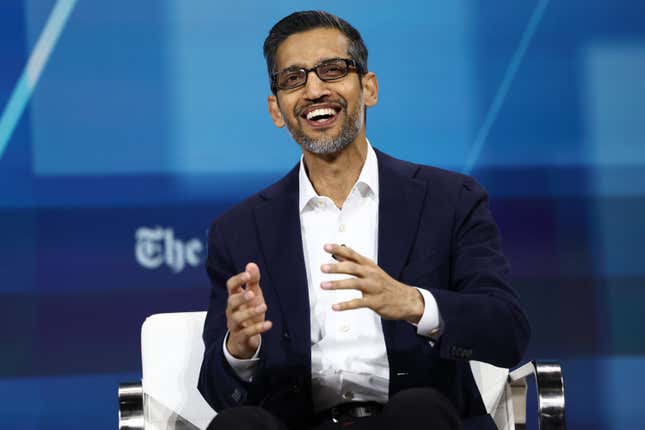 Sundar Pichai smiling while talking with his hands up sitting in a white chair in front of a blue backdrop