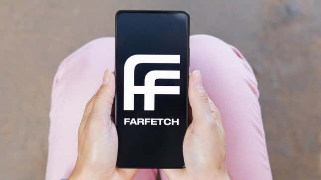 A pair of hands resting on pink pants holds a smartphone with the Farfetch logo displayed.