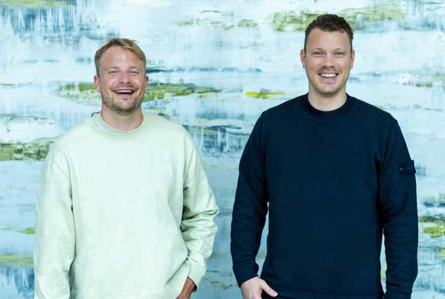 two men stand side by side smiling in front of what looks like a light blue painting with streaks of darker blue and green