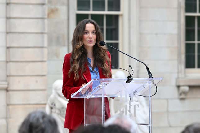 Mira Murati wearing a red suit and blue shirt speaking into a mic at a podium