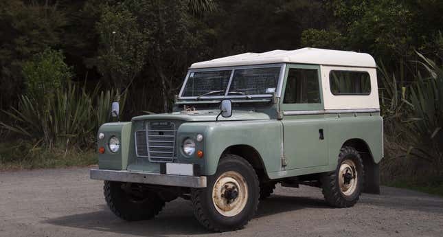 A pale green Land Rover Series III is parked in front of plants