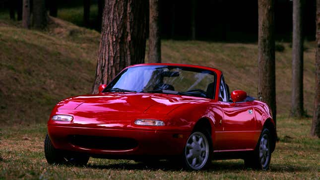 A photo of a red Mazda Miata parked in a forrest. 