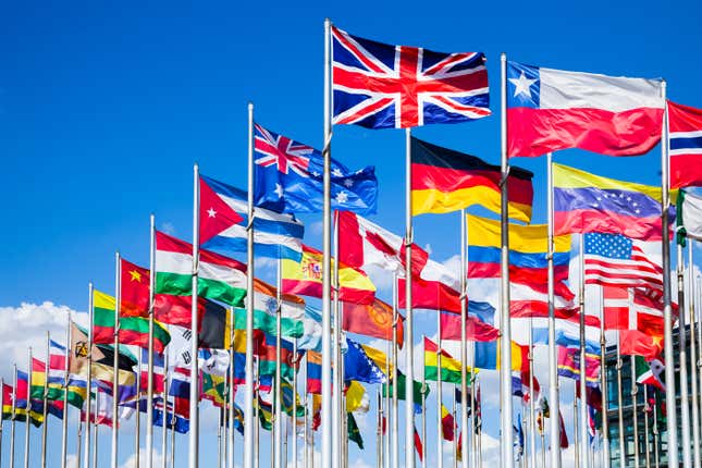 Group of flags of many different nations against blue sky and infront of a convention center.