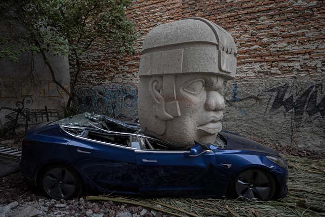 A side view of the blue Tesla Model 3 crushed under the giant Olmec head