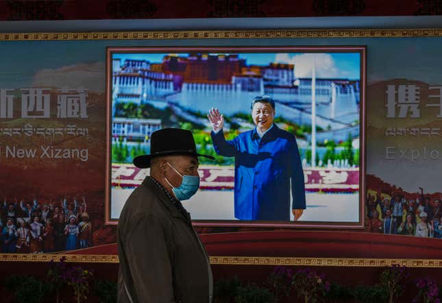 A man in a wide-brimmed hat and covid mask walks in front of a colorful portrait of president Xi waving and smiling. Text in Chinese, Tibetan, and English can be seen on either side reading "New Xizang" ("New Tibet").