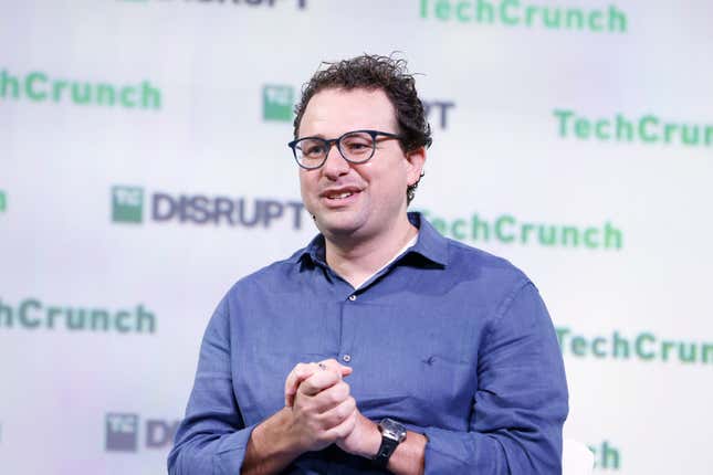 dario amodei speaking with his hands together and wearing a blue button down and glasses in front of a backdrop that says techcrunch disrupt