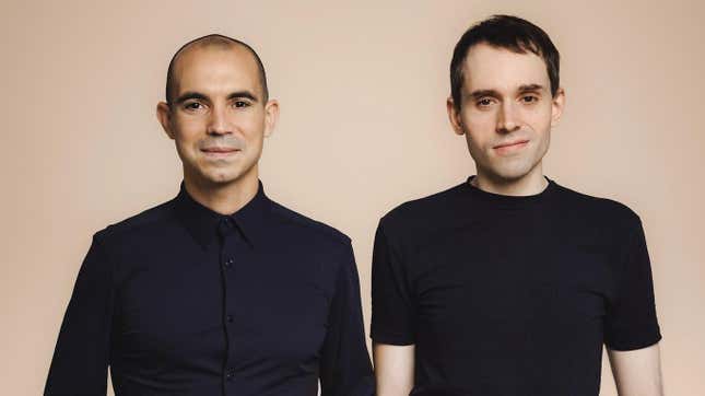 two men standing side by side wearing black shirts in front of a nude color background