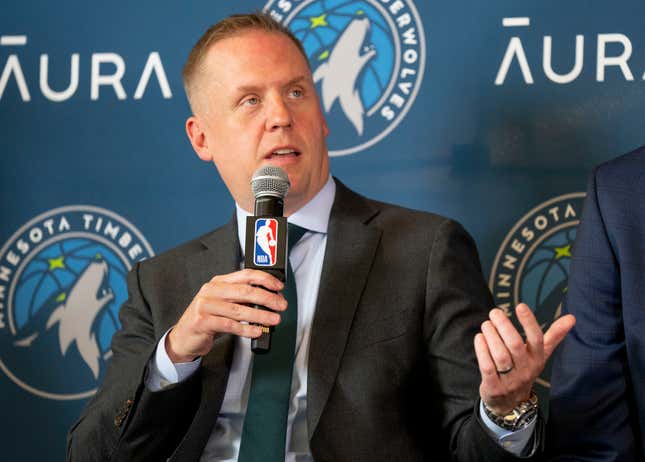 A white man in a black suit speaks into a microphone bearing an NBA logo in front of a Minnesota Timberwolves backdrop.