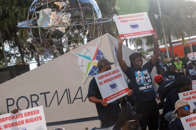 ILA dockworkers in front of a Port of Miami marker hold signs that says "Machines Don't Feed Families" and "Corporate Greed vs. Worker Rights"