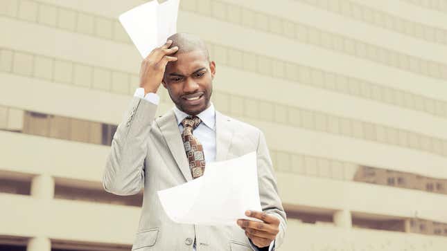 A man in a business suit in front of a building rubs his head in befuddlement at papers he is reading. 