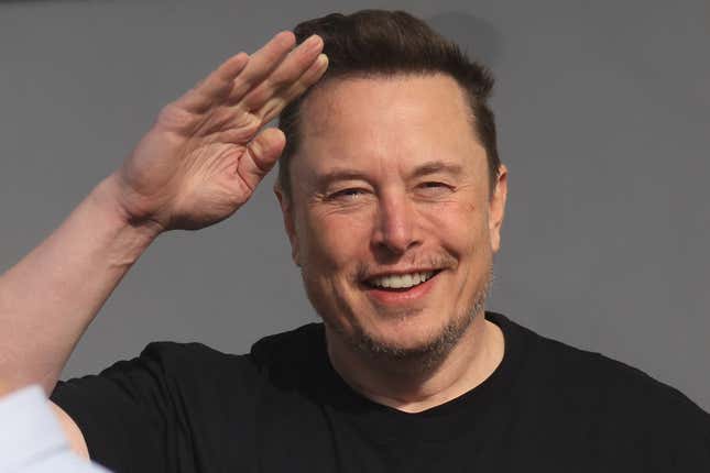 Elon Musk in a black t-shirt raising his hand to block the sun from his eyes, or performing a salute. One of those. 